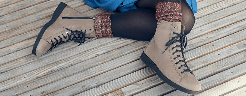 Zwei beige Schnürstiefeletten im Barfußschuh-Stil liegen an den Beinen einer sitzenden Person auf einem Holzboden. Die Stiefeletten haben eine breite Zehenbox, dunkle Schnürsenkel und eine flache, schwarze Sohle. Die Person trägt eine dunkle Hose und gemusterte braune Socken.