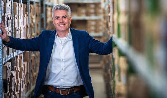 Ein lächelnder Mann in Jeans und Blazer steht in einem Lagergang zwischen Regalen voller Kartons.